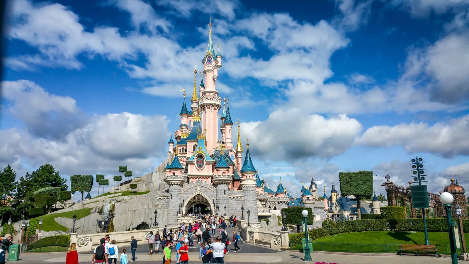 El castell de la Bella Dorment de Disneyland Paris, il·luminat pel sol sota un cel blau amb núvols, amb visitants passejant per l’esplanada.